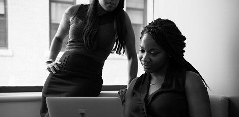 Lady standing next to seated lady both looking at laptop