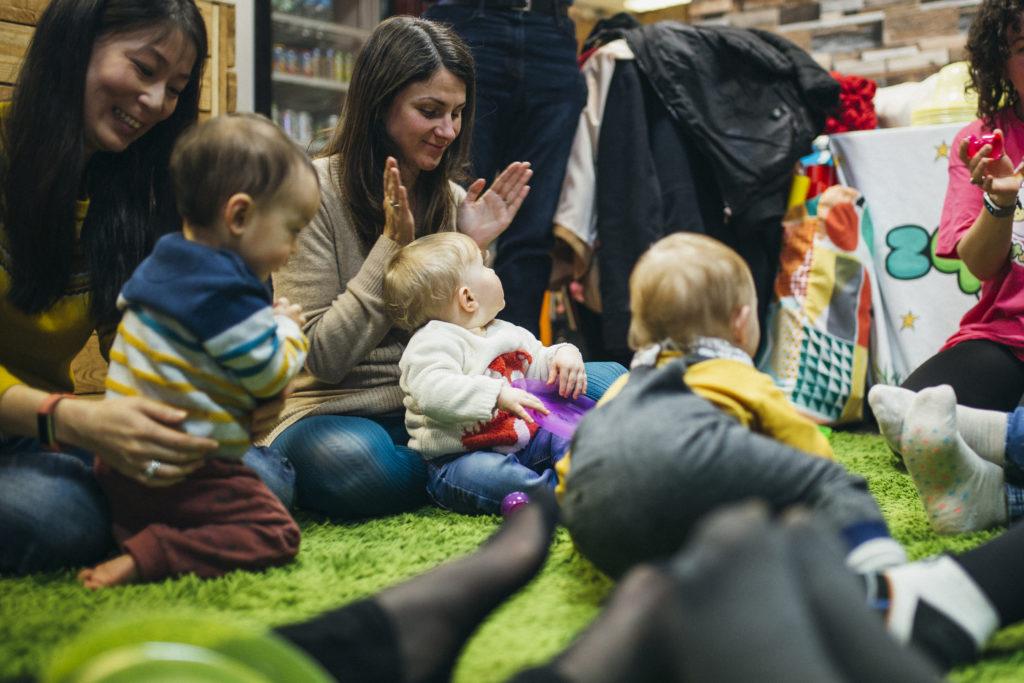 Mothers and their baby children are in a sensory play group. They are singing and dancing together in a circle on the floor.