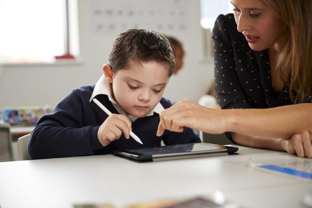 Young female teacher working with a Down syndrome schoolboy sitting at desk using a tablet computer in a primary school classroom, front view, close up